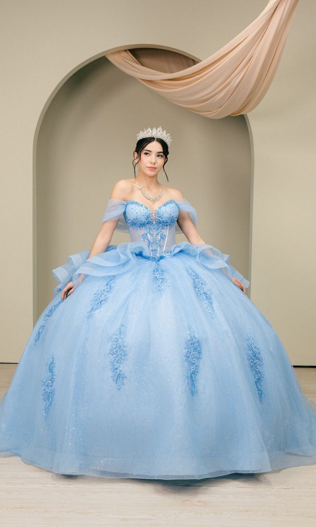 A woman wearing the Quinceanera Dress 8520JQ by Cinderella Couture in blue, featuring a corset bodice and tiara, stands before an arched backdrop with draped fabric, gracefully posing with the glittering skirt spread out.